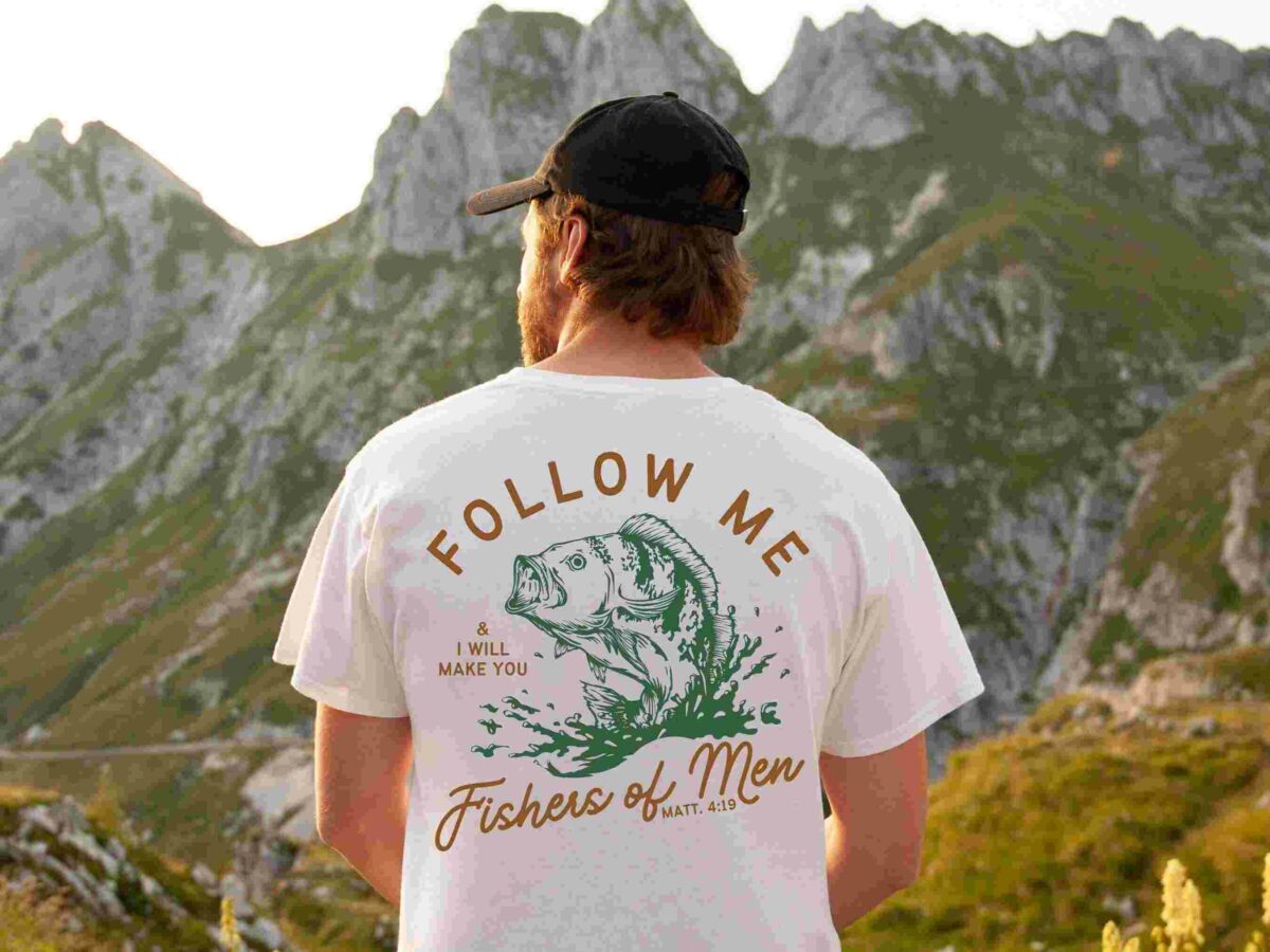 A person wearing a white shirt with a "Follow Me & I Will Make You Fishers of Men" graphic stands facing a mountainous landscape.