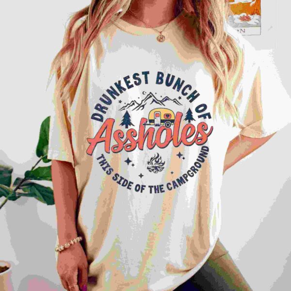 A woman wearing a beige t-shirt with the text "Drunkest bunch of assholes this side of the campground" along with graphics of mountains, trees, a camper, and a campfire. She has long, wavy hair, and stands in front of a plant and a wall decor.