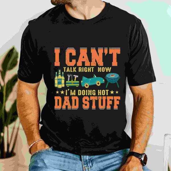 Man wearing a black T-shirt with the text "I can't talk right now, I'm doing hot dad stuff", decorated with images of a drill, a truck, a BBQ grill, and beverages.