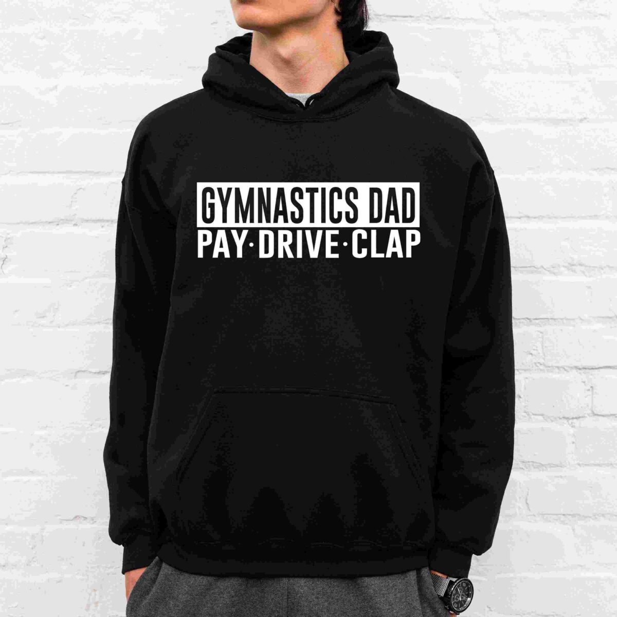 A man wearing a black hoodie stands against a white brick wall. The hoodie has the text "GYMNASTICS DAD" at the top and "PAY. DRIVE. CLAP." below it in bold white letters. The man has his hands in his pockets and is looking to the side.