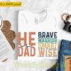 A white T-shirt with the text "He Is Dad" and additional words like "Brave," "Warrior," "Strong," "Smart," "Wise." It is laid out on a white surface near a hat, glasses, a plant, a camera, and a pair of jeans.