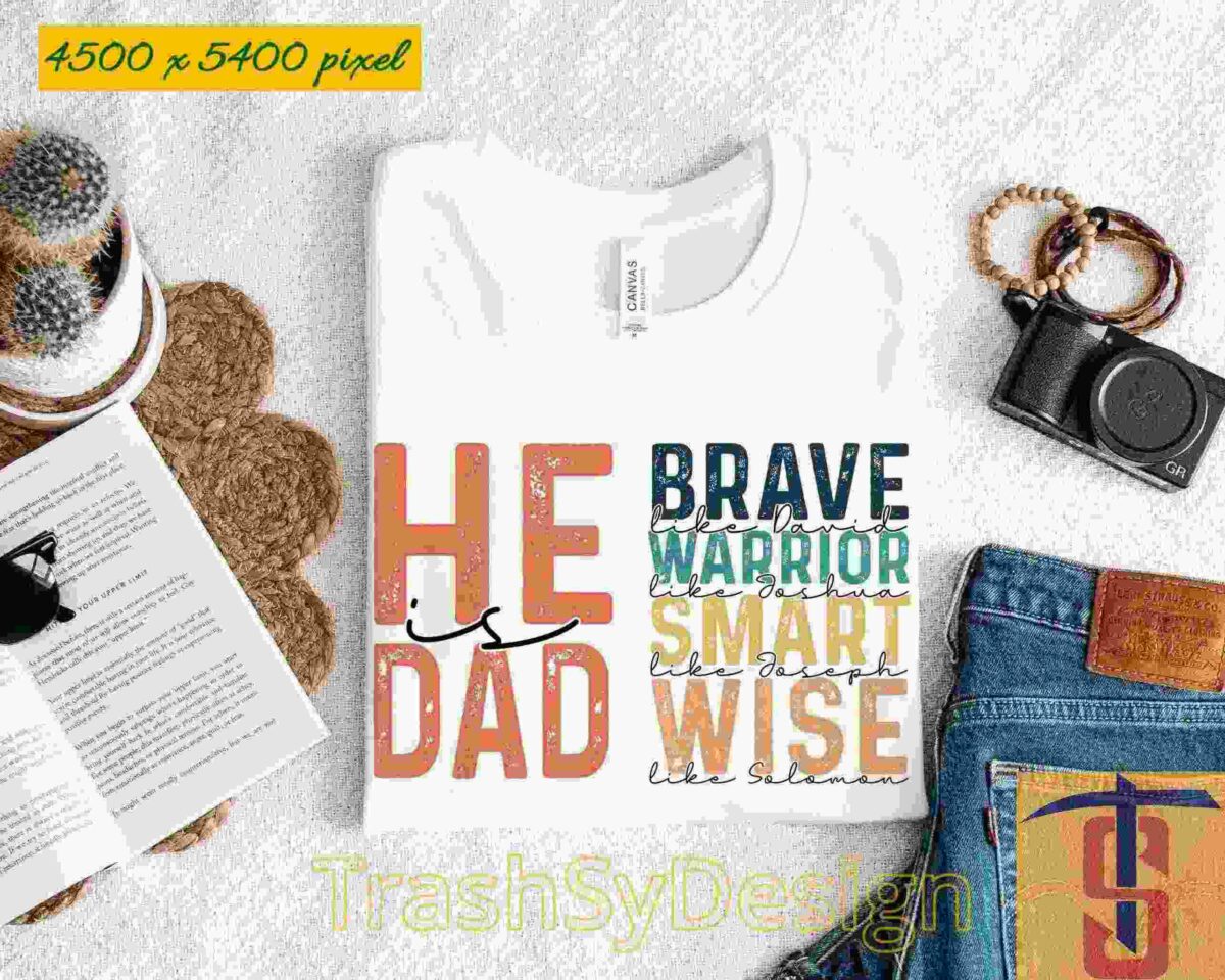 A white T-shirt with the text "He Is Dad" and additional words like "Brave," "Warrior," "Strong," "Smart," "Wise." It is laid out on a white surface near a hat, glasses, a plant, a camera, and a pair of jeans.