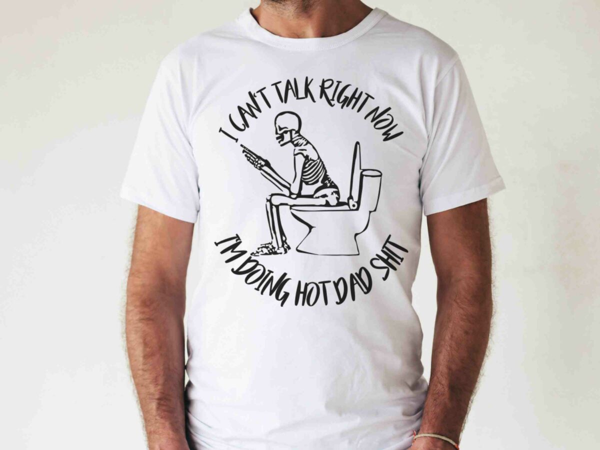 A person wearing a white t-shirt with an illustration of a skeleton sitting on a toilet while using a phone. The text on the shirt reads, "I can't talk right now, I'm doing hot dad shit.