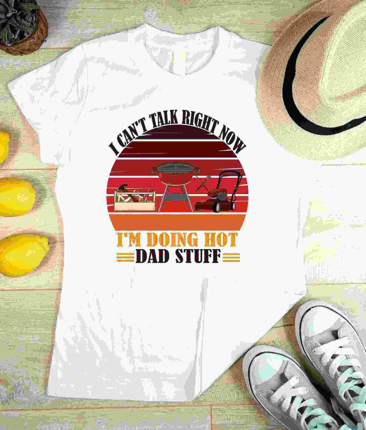 White T-shirt with text "I can't talk right now, I'm doing hot dad stuff" featuring images of a grill, lawn mower, and toolbox. Laid flat beside lemons, a hat, and sneakers on a light wood surface.