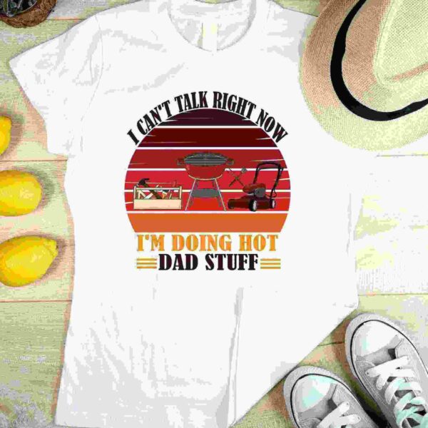 White T-shirt with text "I can't talk right now, I'm doing hot dad stuff" featuring images of a grill, lawn mower, and toolbox. Laid flat beside lemons, a hat, and sneakers on a light wood surface.