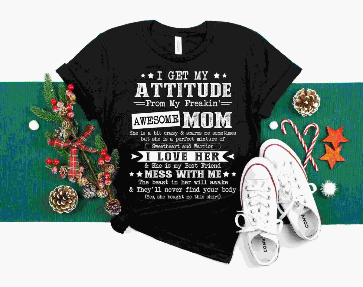 Black t-shirt with white text saying: "I get my attitude from my freakin' awesome mom. She is a bit crazy & scares me sometimes but she is a perfect mixture of sweetheart & warrior." Surrounded by holiday decorations.