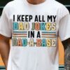 A person is wearing a white t-shirt with the text "I KEEP ALL MY DAD JOKES IN A DAD-A-BASE" in bold, colorful, and stylized letters. The person has a tattoo on their forearm.