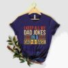A dark gray t-shirt hangs on a wooden hanger against a neutral background. The shirt features colorful text that reads, "I keep all my dad jokes in a dad-a-base." A small branch of green leaves is pinned to the bottom of the shirt.