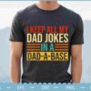A person wearing a dark t-shirt with the text: "I keep all my dad jokes in a dad-a-base." The background is out of focus, showing an outdoor setting.