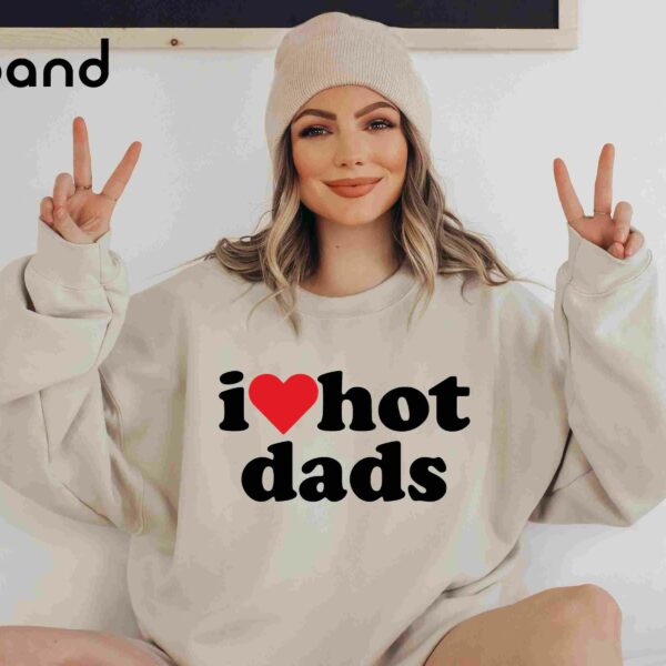 A person wearing a beige beanie and an oversized sweatshirt that reads "i ♥ hot dads" with both hands raised, displaying peace signs. The word "Sand" is in the top left corner.