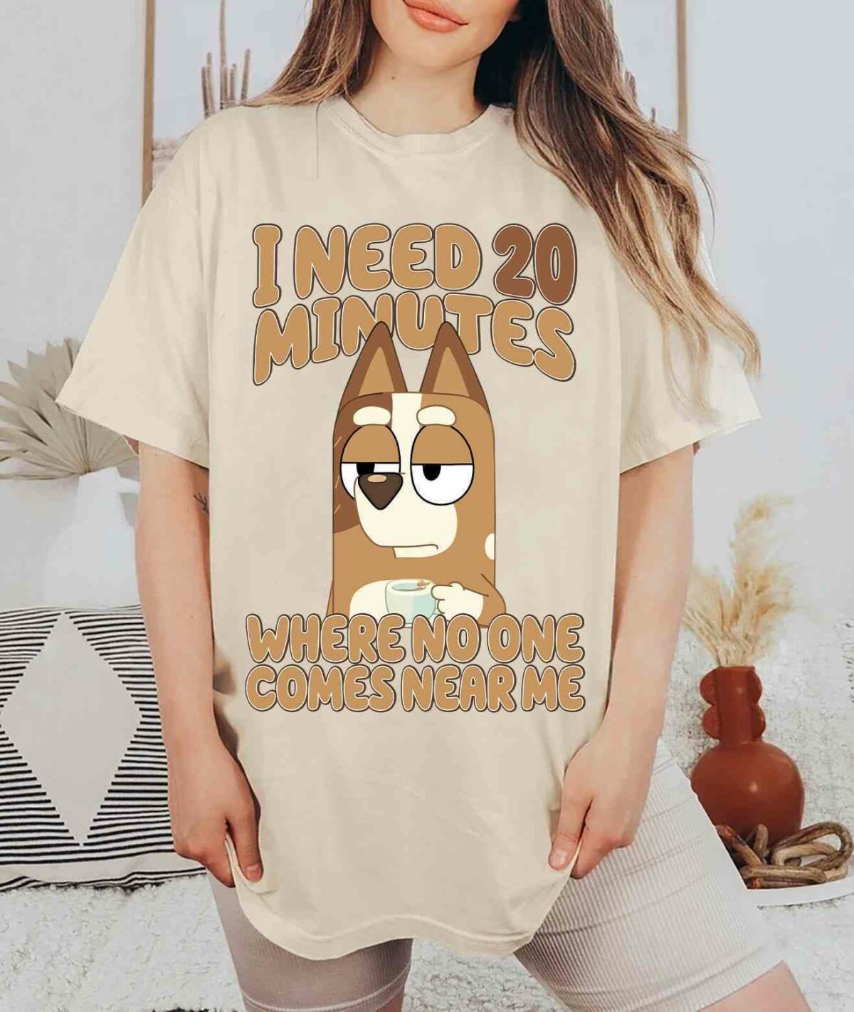 Person wearing a beige T-shirt with an illustration of a dog holding a mug and the text, "I need 20 minutes where no one comes near me." The background shows a cozy room with a bed and cushions.