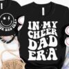 Two black shirts with white text. One says "IN MY CHEER DAD ERA" and the other has a smiley face with "CHEER DAD ERA." Nearby are white sneakers, a rope, and a black and white patterned bag.