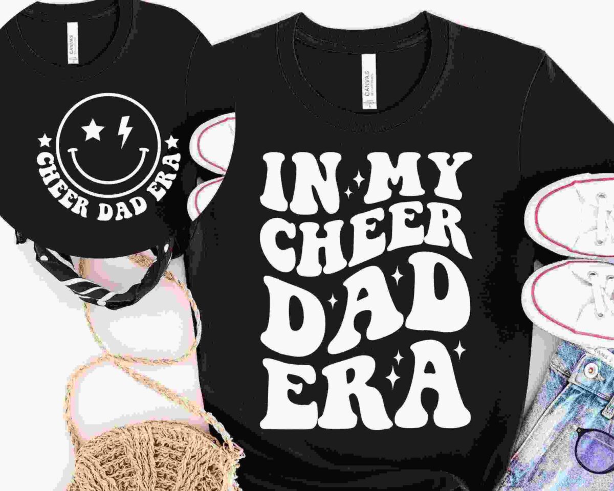 Two black shirts with white text. One says "IN MY CHEER DAD ERA" and the other has a smiley face with "CHEER DAD ERA." Nearby are white sneakers, a rope, and a black and white patterned bag.