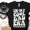 Black T-shirt with the text "IN MY COOL DAD ERA" in bold, playful font, surrounded by a bandana, white sneakers, denim shorts, a straw hat, and a matching black round bag with "COOL DAD ERA" and smiley face.