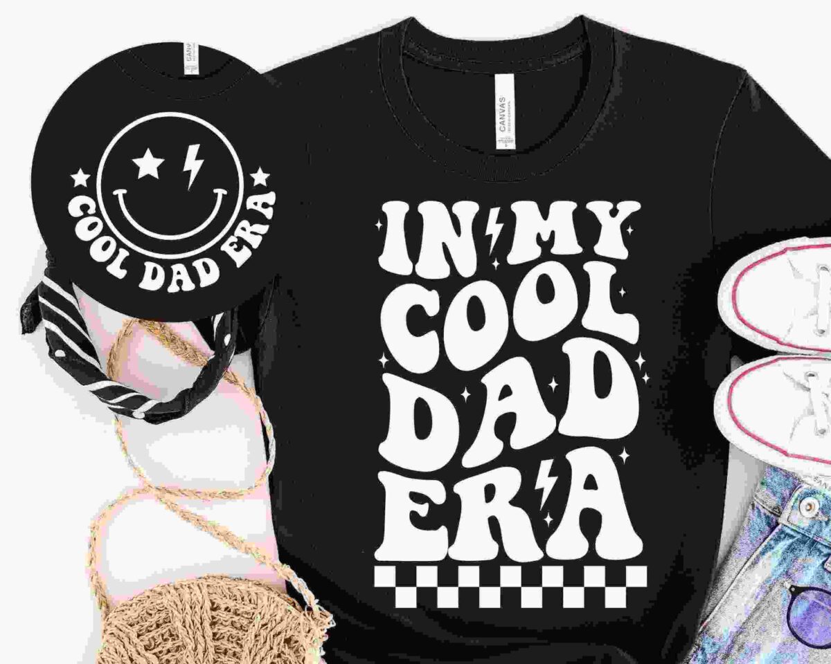 Black T-shirt with the text "IN MY COOL DAD ERA" in bold, playful font, surrounded by a bandana, white sneakers, denim shorts, a straw hat, and a matching black round bag with "COOL DAD ERA" and smiley face.