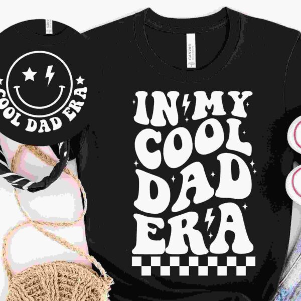 Black T-shirt with the text "IN MY COOL DAD ERA" in bold, playful font, surrounded by a bandana, white sneakers, denim shorts, a straw hat, and a matching black round bag with "COOL DAD ERA" and smiley face.