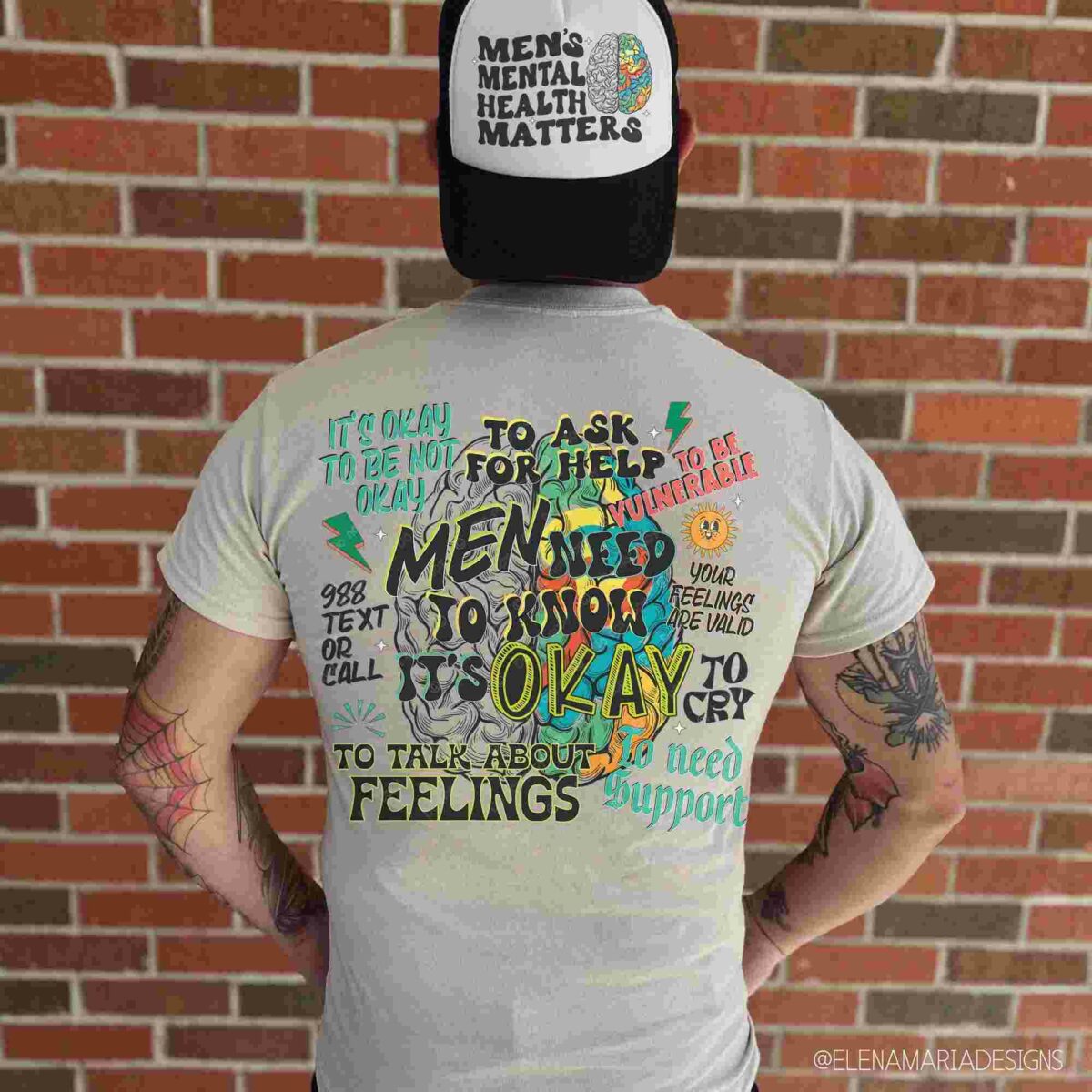 A person wearing a hat and t-shirt with messages promoting men's mental health stands against a brick wall. The t-shirt features phrases encouraging open discussion about men's feelings.