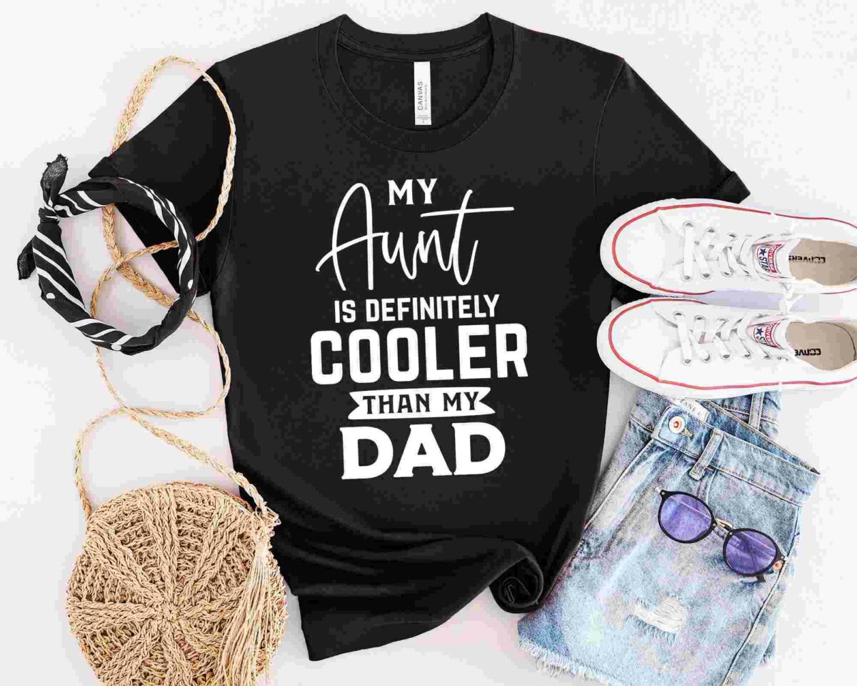 A black t-shirt with the text "My Aunt is Definitely Cooler Than My Dad" displayed alongside a pair of white sneakers, a woven bag, a black and white scarf, denim shorts, and purple sunglasses.