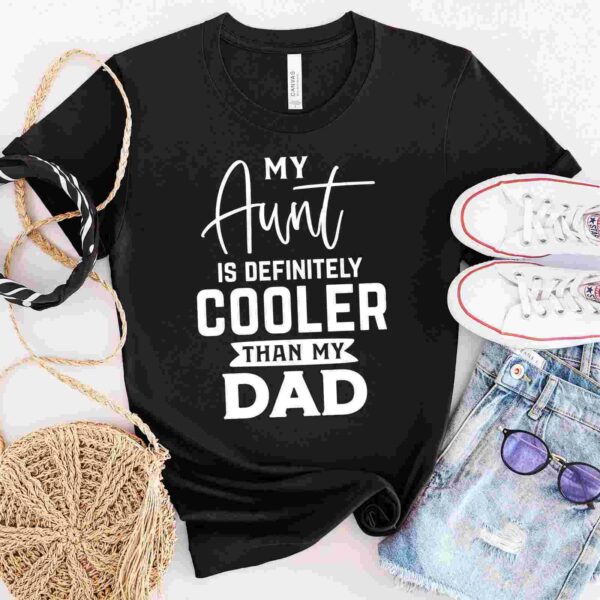 A black t-shirt with the text "My Aunt is Definitely Cooler Than My Dad" displayed alongside a pair of white sneakers, a woven bag, a black and white scarf, denim shorts, and purple sunglasses.