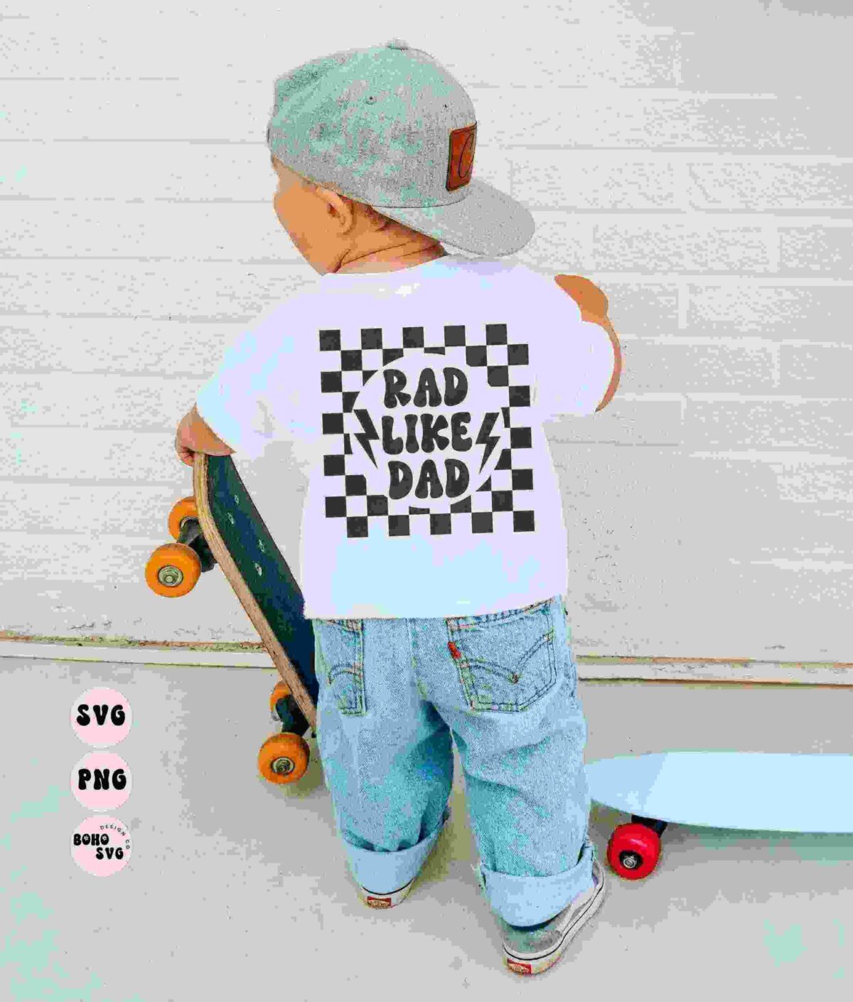 A young child in a grey hat and white T-shirt with "Rad Like Dad" text stands on a skateboard while holding another skateboard. The child faces away from the camera against a white brick wall.