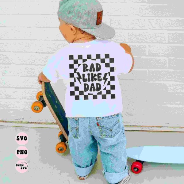 A young child in a grey hat and white T-shirt with "Rad Like Dad" text stands on a skateboard while holding another skateboard. The child faces away from the camera against a white brick wall.