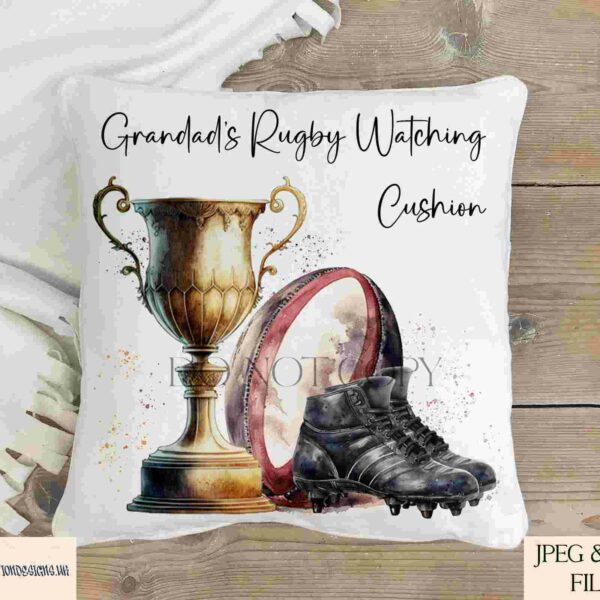 A throw pillow featuring a design with a trophy, a rugby ball, and cleats. Text reads "Grandad's Rugby Watching Cushion." The pillow is placed on a wooden surface with a white fabric in the background.