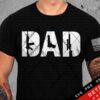 Alt Text: A person wears a black T-shirt with the word "DAD" in large letters, featuring a design with firearm silhouettes, and an American flag patch on the right sleeve. Tattoos are visible on both arms.