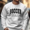 A man wearing a white sweatshirt that says "SOCCER DAD" in large black letters is walking on a city street.