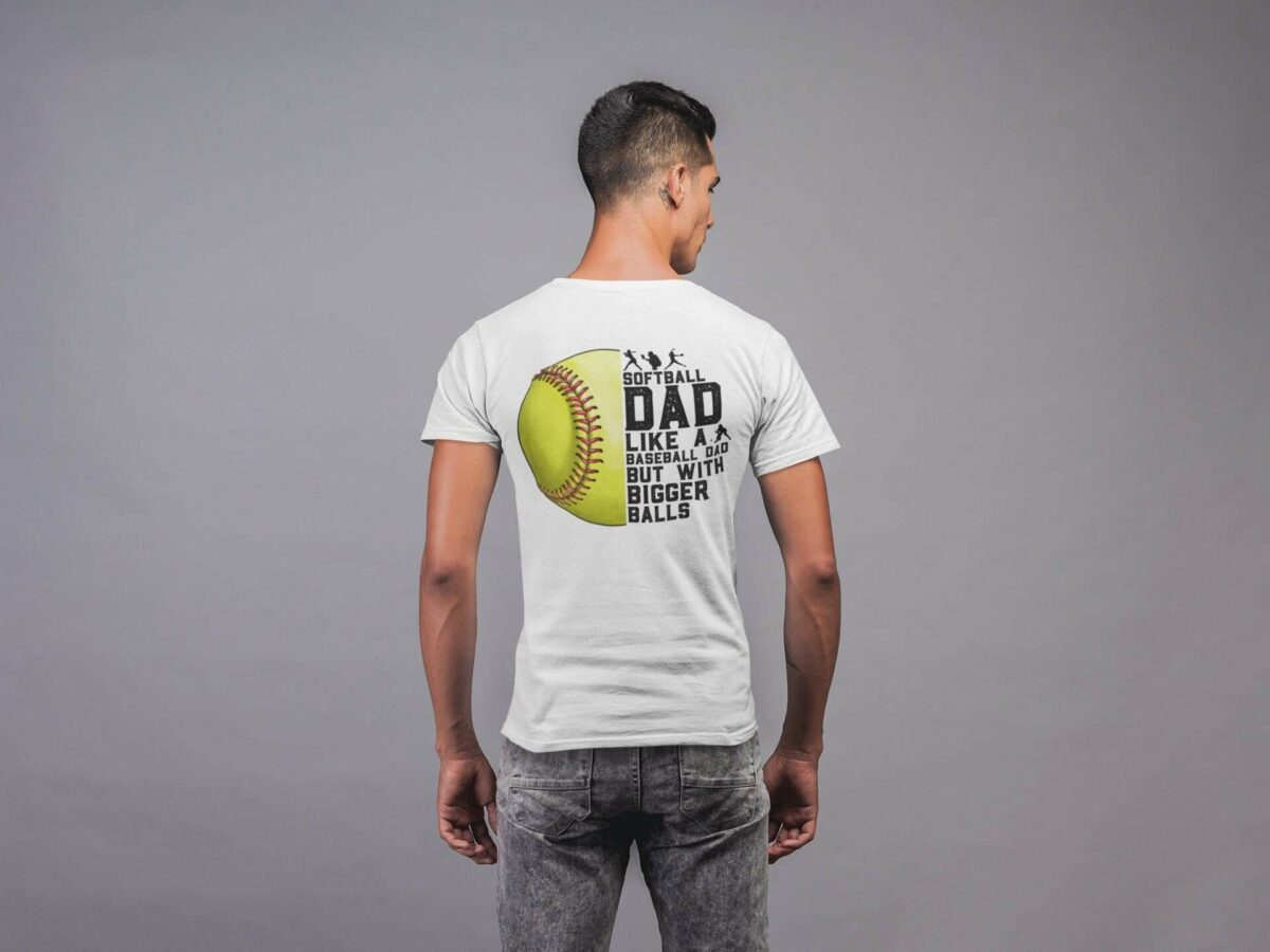 A person stands facing away, wearing a white shirt with a graphic showing a softball and text that reads, "Softball Dad: Like a Baseball Dad but With Bigger Balls.