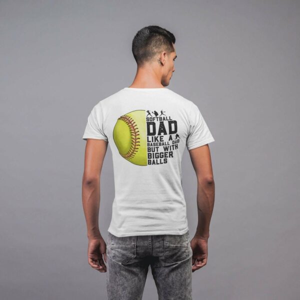 A person stands facing away, wearing a white shirt with a graphic showing a softball and text that reads, "Softball Dad: Like a Baseball Dad but With Bigger Balls.