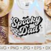 A white sweatshirt with a "Spooky Dad" design featuring a bat and spider web, displayed beside a straw hat and beads.