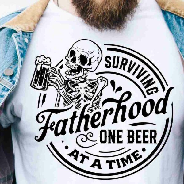 A person wearing a white t-shirt under a denim jacket. The t-shirt features a graphic of a skeleton holding a beer mug and reads "Surviving Fatherhood One Beer At a Time" in bold, stylized font.