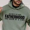 A man wears a green hoodie with the text "Surviving Fatherhood One Beer at a Time." The image includes a watermark that reads "SVG Mode.