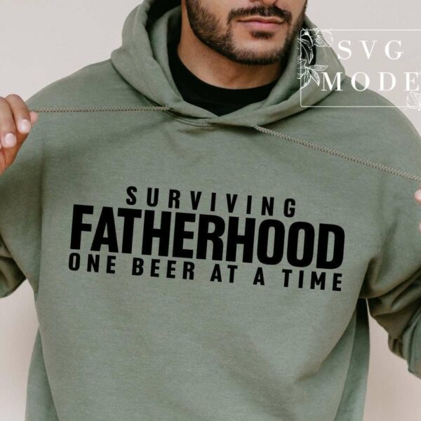 A man wears a green hoodie with the text "Surviving Fatherhood One Beer at a Time." The image includes a watermark that reads "SVG Mode.