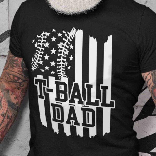 Close-up of a man's torso with tattoos wearing a black shirt that reads "T-Ball Dad" with a baseball-stitched American flag design.