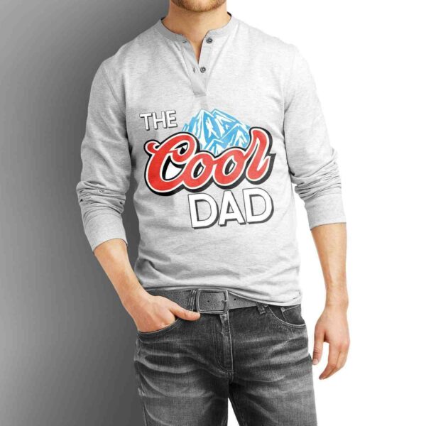 A person is wearing a light gray long-sleeve henley shirt with the text "The Cool Dad" and a graphic of a mountain in blue. The person is standing with one hand in the pocket of their dark jeans. The background is a gradient from white to dark gray.