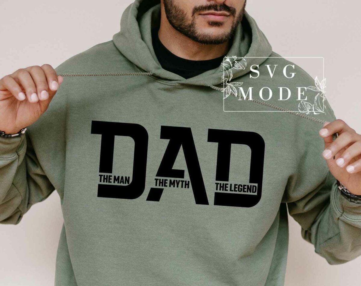 A person wearing a green hoodie that has the word "DAD" printed in large black letters on the front, with smaller text underneath that reads "THE MAN, THE MYTH, THE LEGEND." The individual is pulling the hoodie strings with both hands.