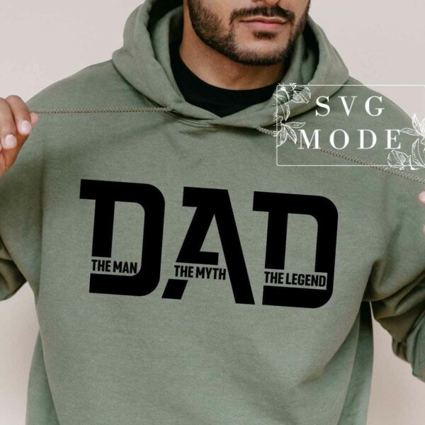 A person wearing a green hoodie that has the word "DAD" printed in large black letters on the front, with smaller text underneath that reads "THE MAN, THE MYTH, THE LEGEND." The individual is pulling the hoodie strings with both hands.