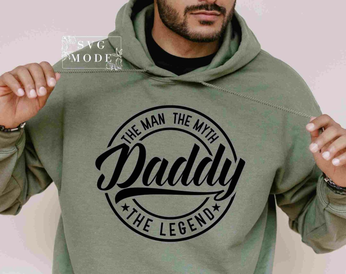 A person wears a green hoodie featuring black text that reads "Daddy, The Man, The Myth, The Legend." The person is pulling on the hoodie strings.