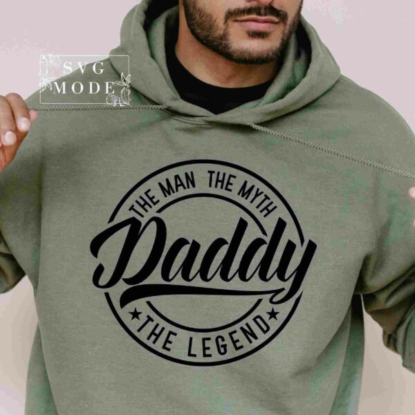 A person wears a green hoodie featuring black text that reads "Daddy, The Man, The Myth, The Legend." The person is pulling on the hoodie strings.