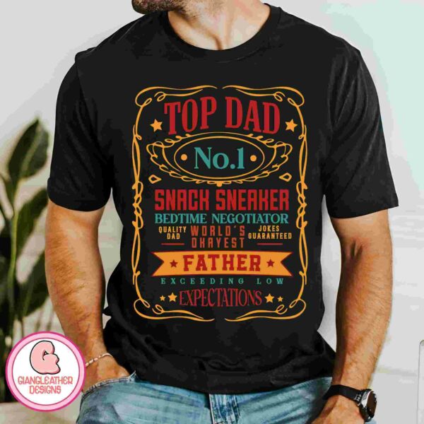 A person wearing a black shirt with colorful text that reads "Top Dad No.1, Snack Sneaker, Bedtime Negotiator, World's Okayest Dad, Quality Jokes, Father Exceeding Low Expectations.