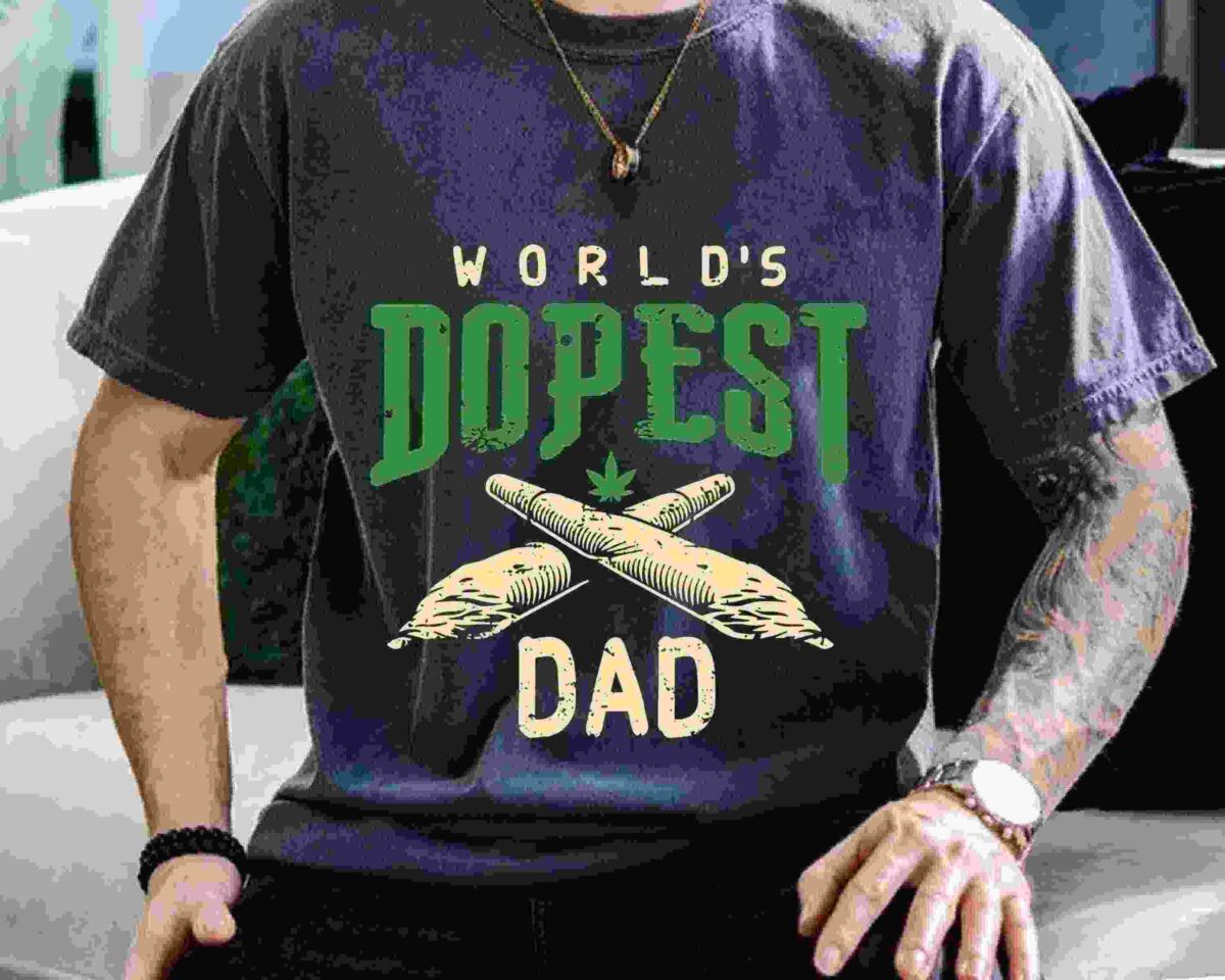 A person wearing a gray T-shirt with the text "World's Dopest Dad" featuring two crossed rolled items and a small cannabis leaf.