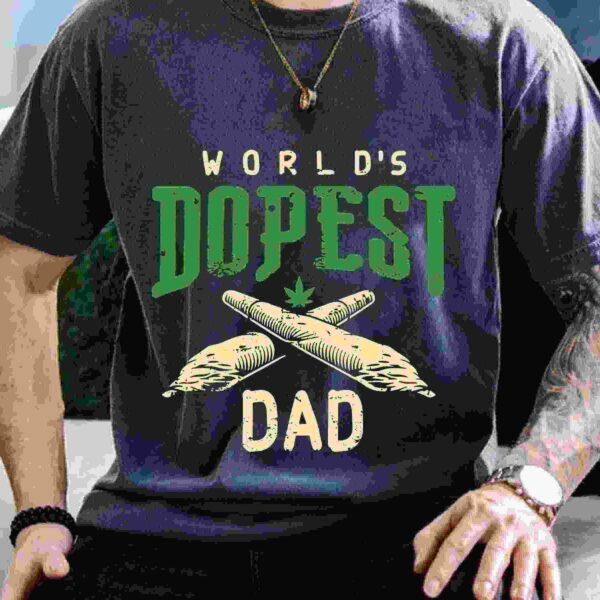 A person wearing a gray T-shirt with the text "World's Dopest Dad" featuring two crossed rolled items and a small cannabis leaf.